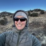 Woman out hiking while on a glamping trip wearing a hat, coat and standing on a hill slope.