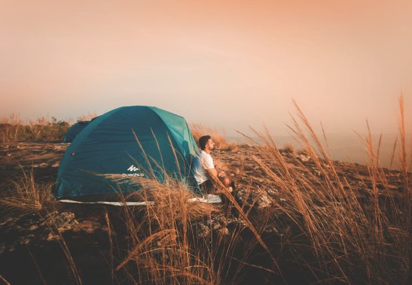 A man enjoys a peaceful glamping experience beside a blue tent in a tranquil natural setting during sunset.